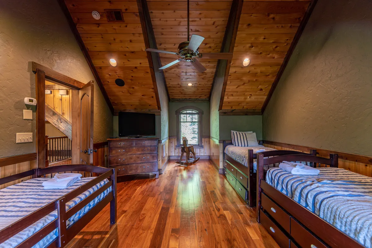 A bright bedroom with a wooden ceiling, twin beds, and a dresser, illuminated by a window with scenic views. The arrangement hints at a playful and welcoming space, ideal for children or multiple guests to rest, read, or play games together.