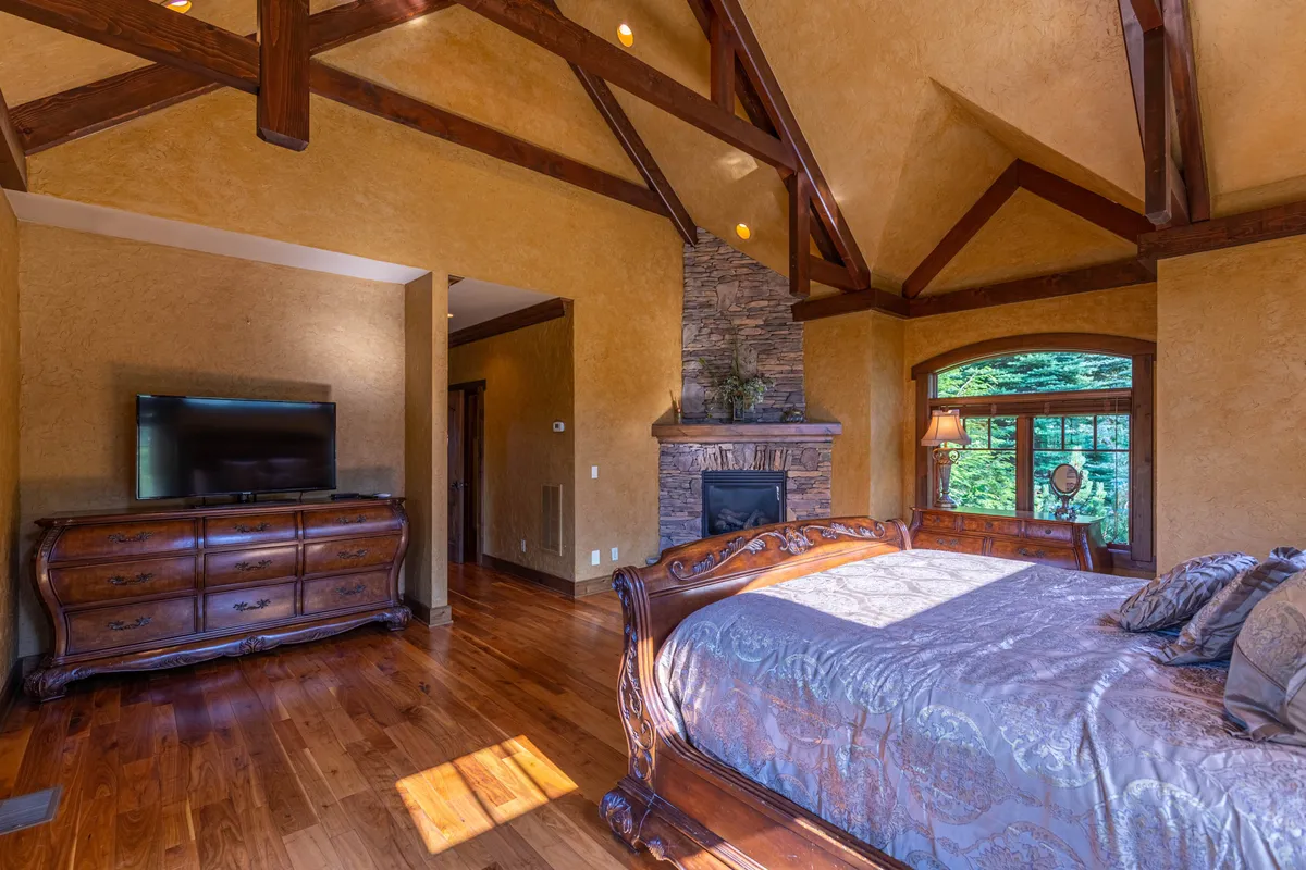 This inviting bedroom features a king-sized bed, a stone fireplace, and a large window that floods the space with natural light. The wooden furniture and warm tones create a tranquil retreat, perfect for couples seeking a relaxing sanctuary. The window offers views of the outdoors, and the fireplace adds a cozy touch for peaceful evenings.