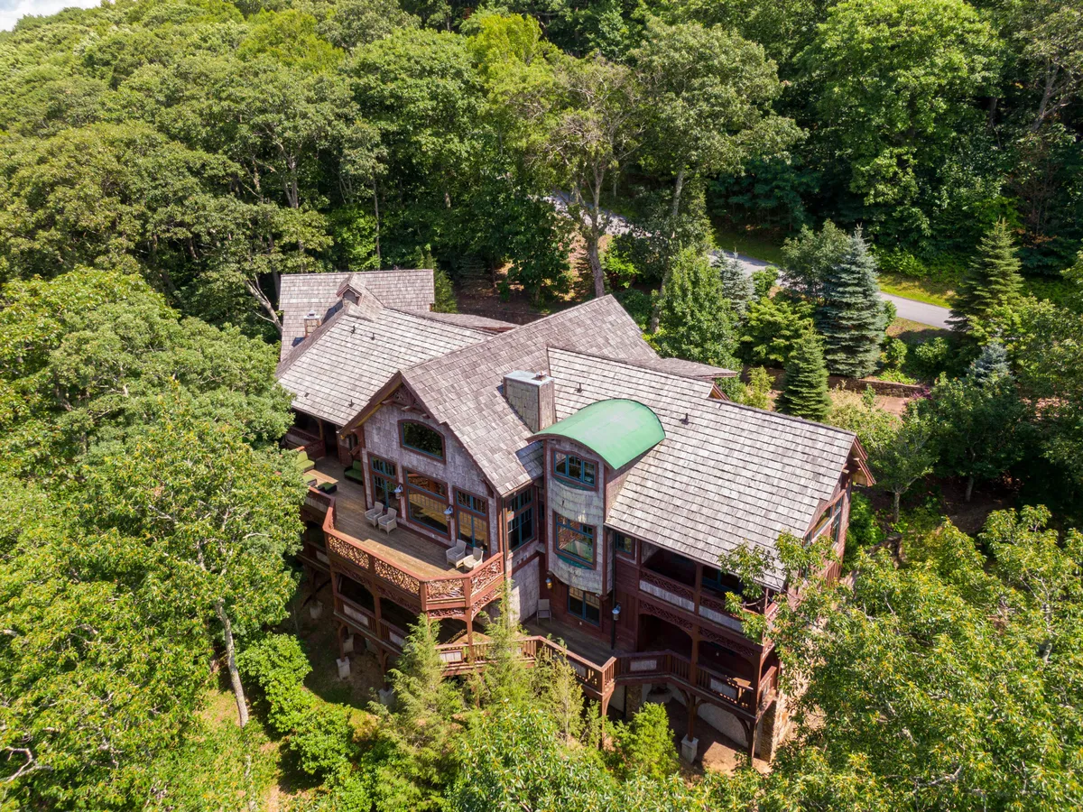 A wide exterior view of the property features the large, multi-gabled roof, wooden balcony, expansive windows, and lush surrounding greenery. The scene evokes a tranquil connection to nature and highlights inviting outdoor spaces, suggesting opportunities for outdoor gatherings and relaxation.