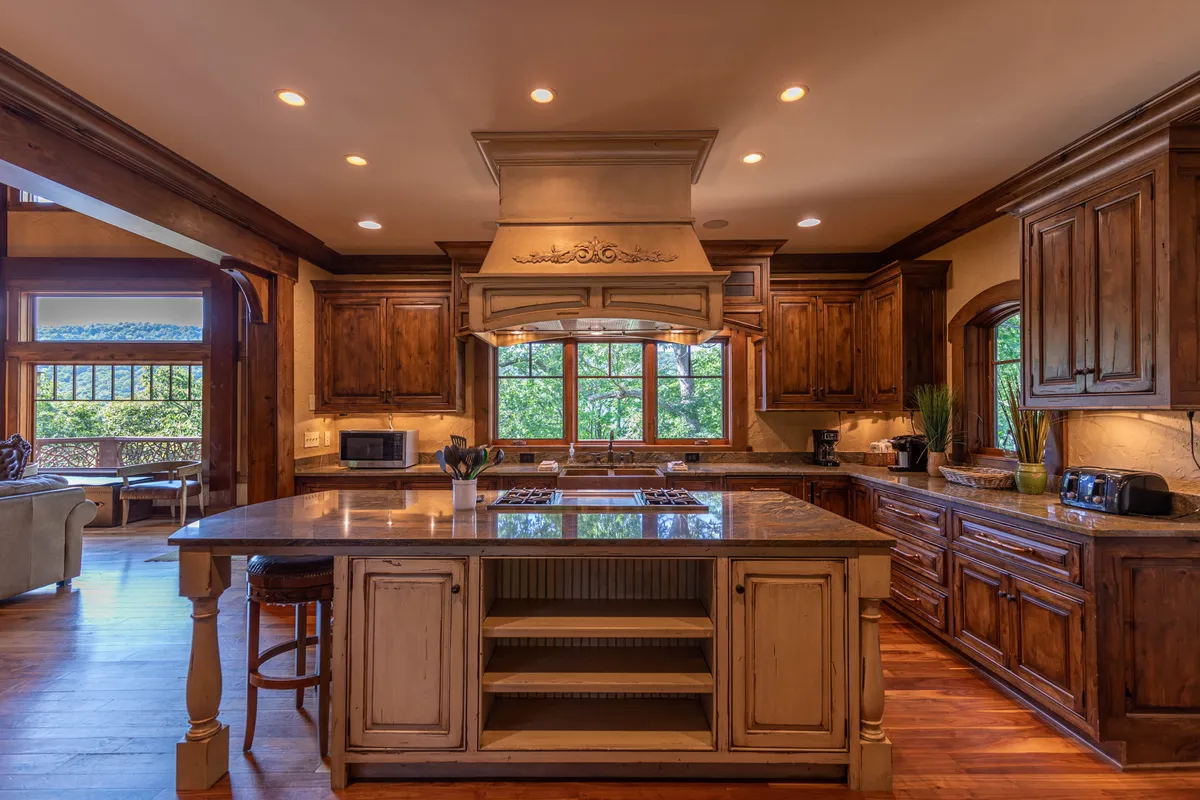 A warmly lit kitchen featuring a large central island with granite countertops, custom wooden cabinetry, and a window offering views to the outdoors. The space is designed for gathering and meal prep, with ample room for multiple people to cook or socialize.