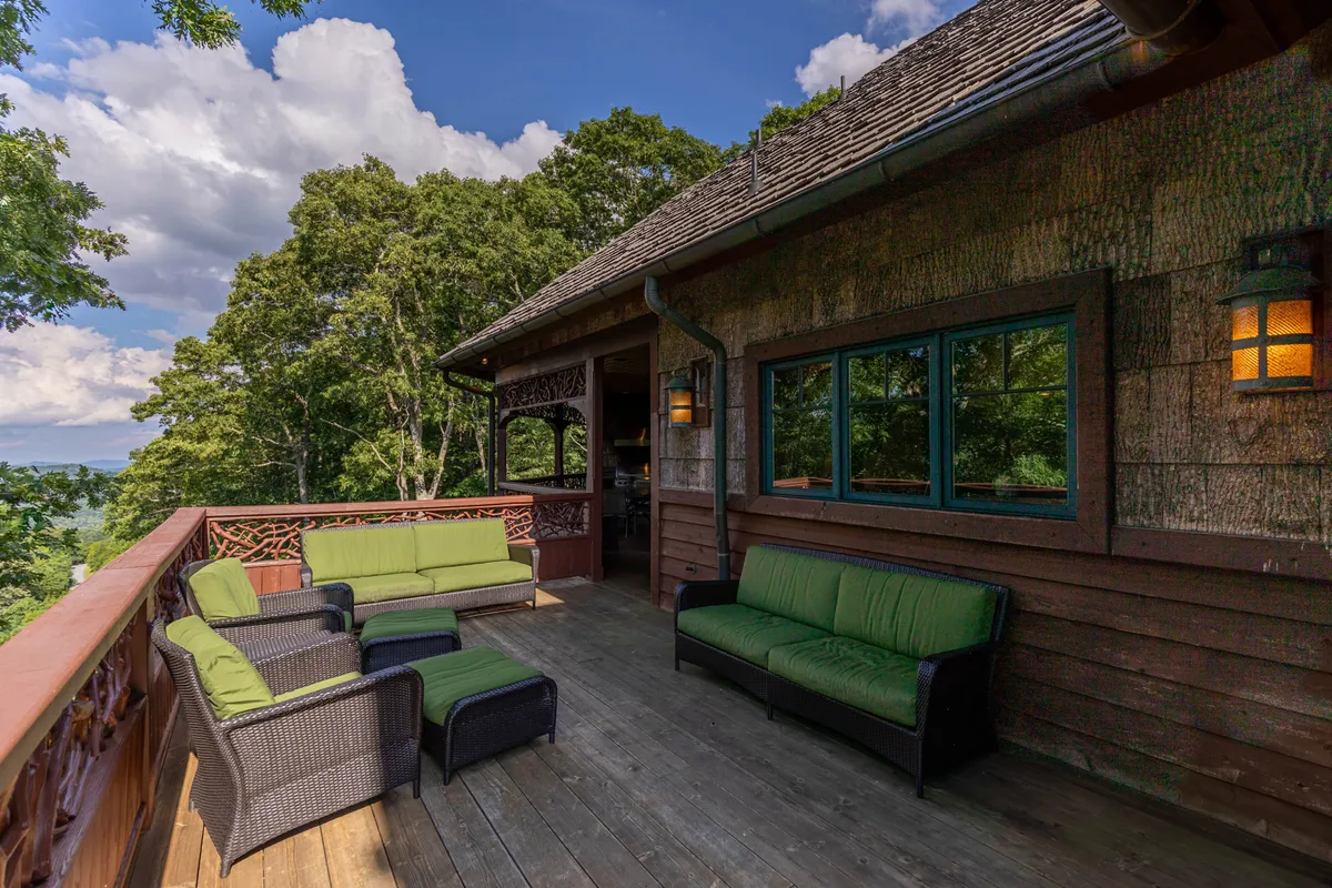 A tranquil exterior shot of a spacious wooden deck surrounded by lush trees. The deck features a green cushioned seating area perfect for group conversations or outdoor games, and offers ample space for gathering and enjoying fresh mountain air.