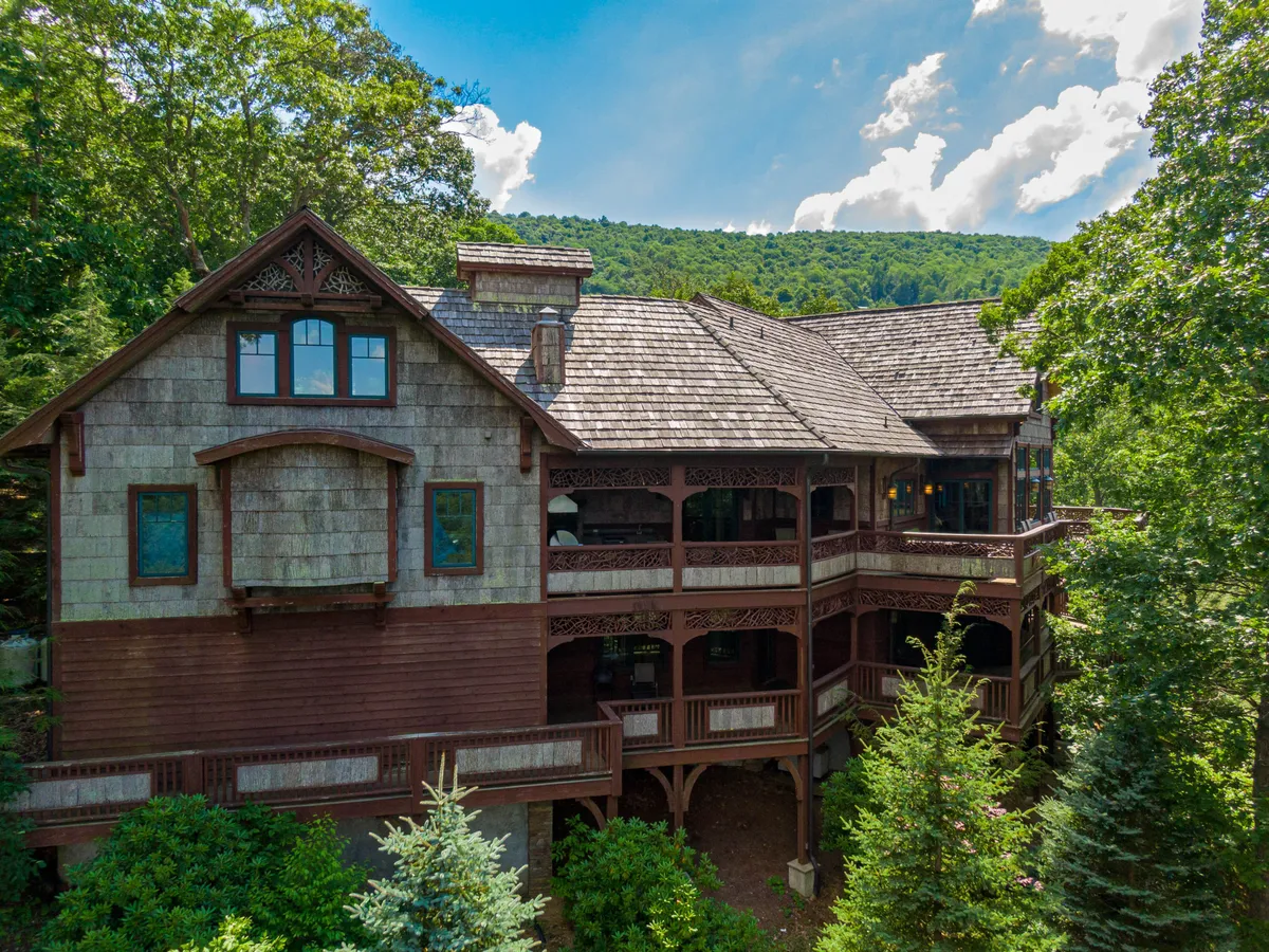 The image showcases the striking exterior of a luxury single-family home, featuring beautifully crafted wooden balconies, distinctive shingled roofing, and surrounded by vibrant, lush greenery. The setting suggests tranquil moments spent on the balconies with panoramic mountain views and intimate gatherings beneath the trees as the sun sets.