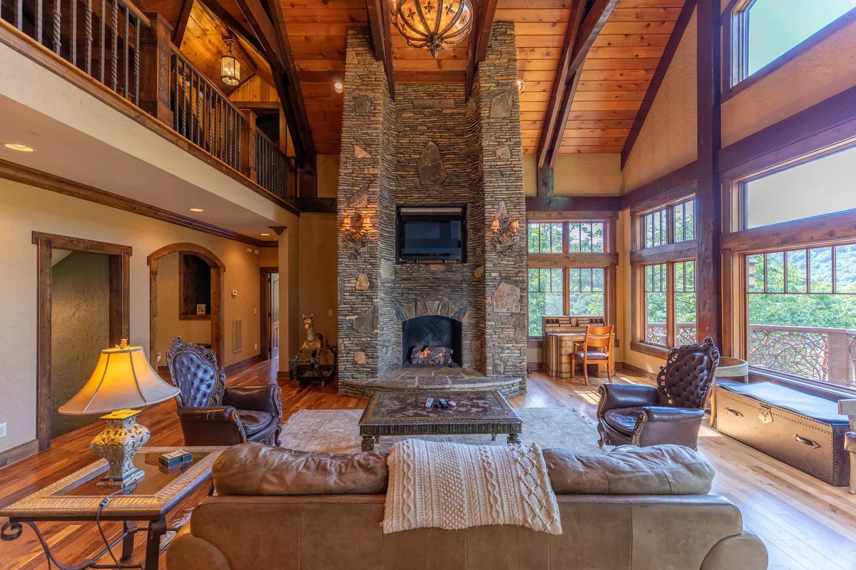 This image captures a sunlit great room with exposed wooden beams, a dramatic stone fireplace, large windows revealing mountain views, and plush seating arranged for conversation or relaxation. The ambiance is cozy and inviting, with natural light enhancing the artisanal craftsmanship.