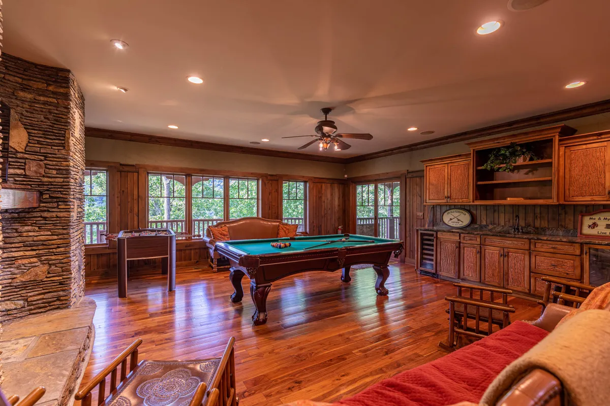 A warm and casual game room interior featuring a pool table, a stone fireplace, wooden cabinetry, and large windows with natural light. People are gathered playing billiards, socializing, and relaxing by the fire.