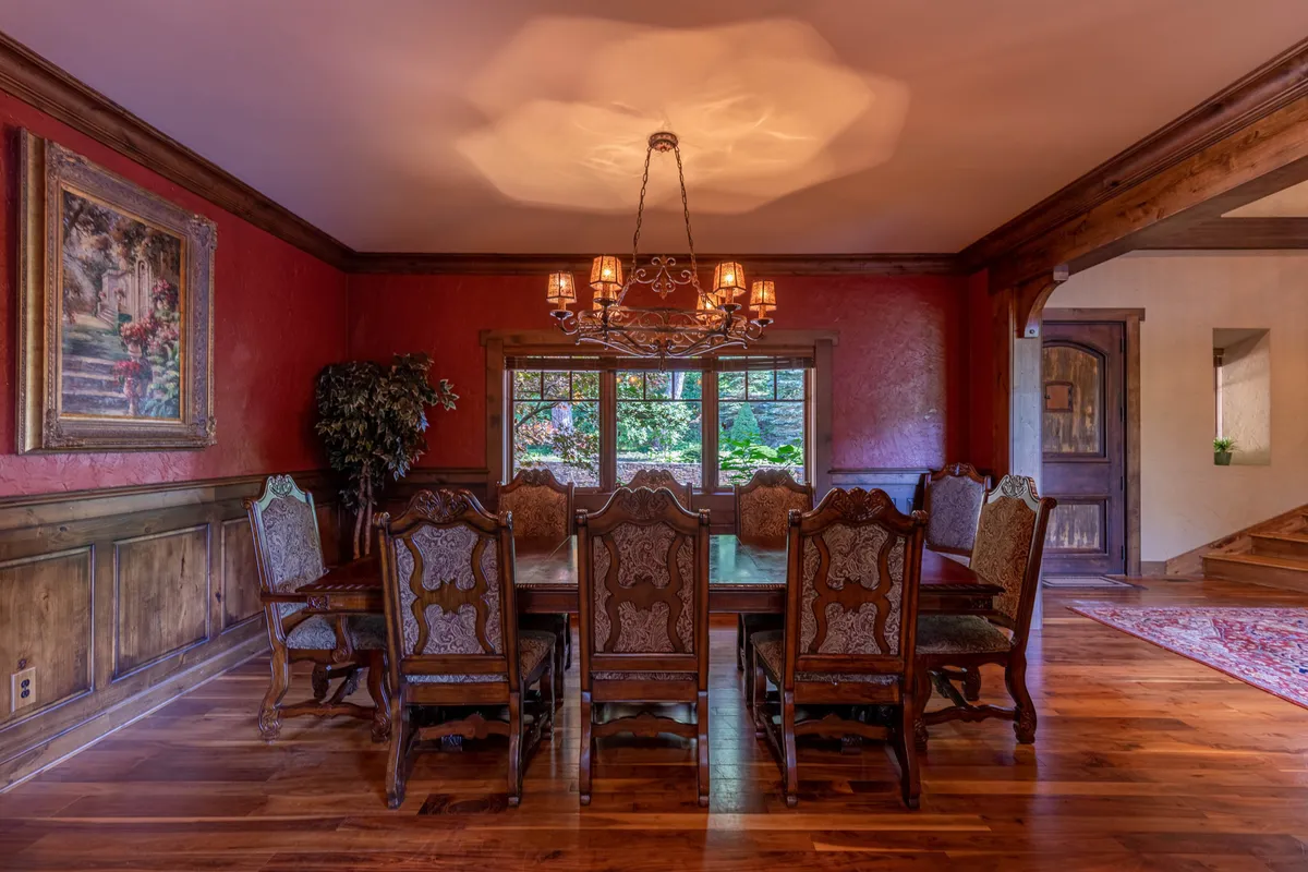A large wooden dining table sits beneath an ornate chandelier, with framed artwork adorning the wall and expansive windows looking into lush gardens. The setting is elegant yet cozy—perfect for shared meals and celebrations.