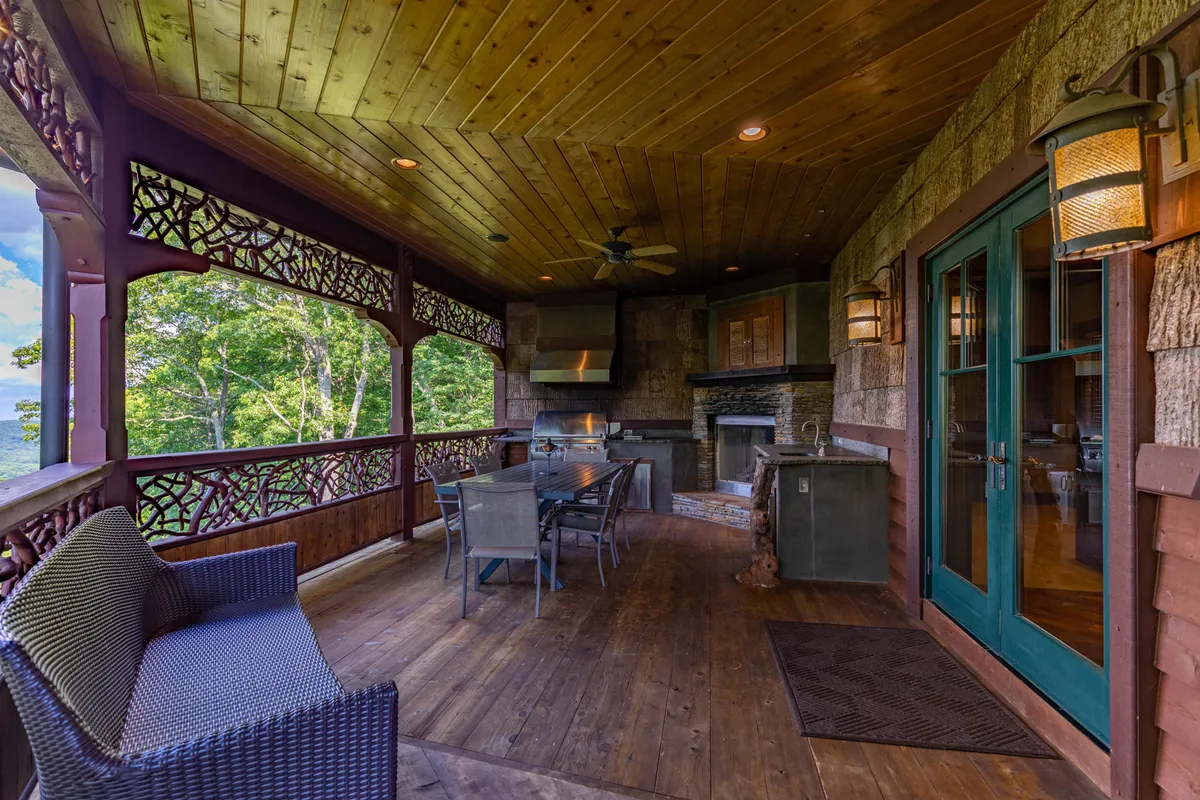 This image showcases a cozy outdoor dining table surrounded by comfortable seating and a stone fireplace, all under the shelter of a covered deck. The intricate wooden railing and the overall design signal luxury outdoor living, perfect for year-round enjoyment.